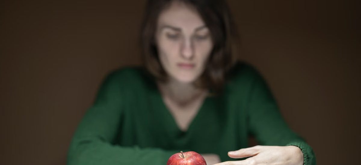 A woman in green thoughtfully observes a red apple on a white table indoors.