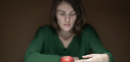 A woman in green thoughtfully observes a red apple on a white table indoors.