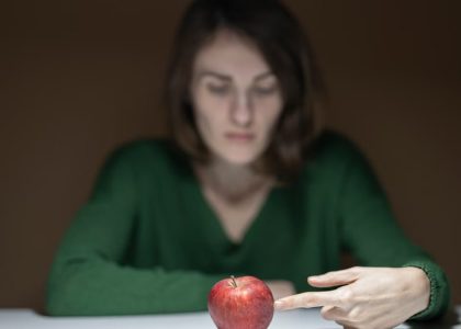 A woman in green thoughtfully observes a red apple on a white table indoors.